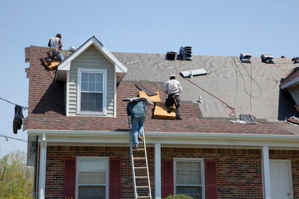 Roofers replace shingles on a two-story house, standing on the roof with a ladder and tools under a clear blue sky.